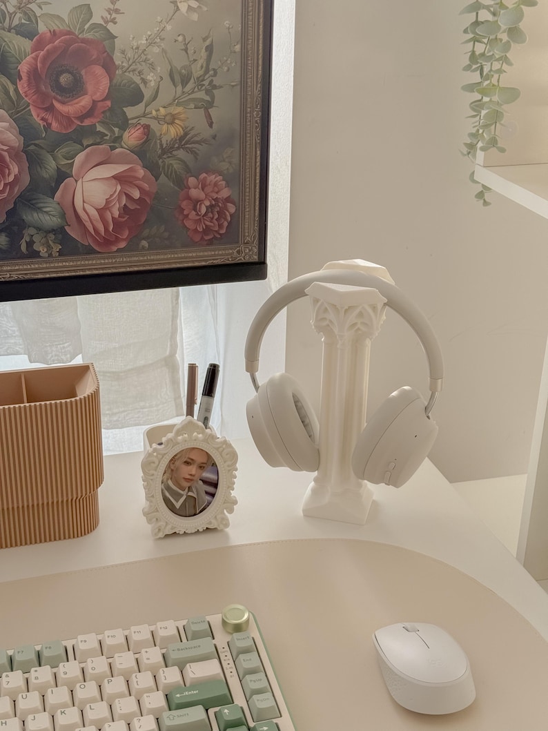 May include: A white desk setup featuring a decorative headphone stand, a framed photo, and a keyboard with mint green keys. A pair of white headphones rests on the stand. A floral painting hangs in the background.