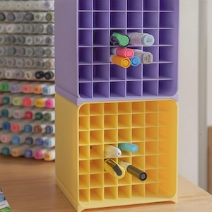 May include: A stack of two square plastic storage organizers with a grid of square compartments. The top unit is light purple and the bottom unit is yellow. Several markers in various colors are inserted into the individual grid slots.