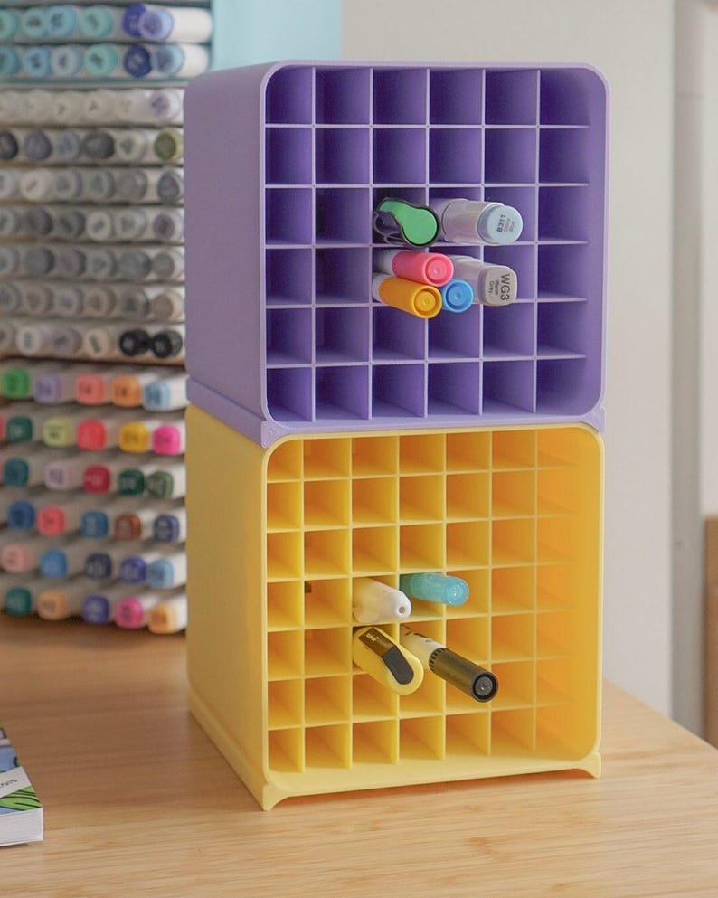 May include: A stack of two square plastic storage organizers with a grid of square compartments. The top unit is light purple and the bottom unit is yellow. Several markers in various colors are inserted into the individual grid slots.