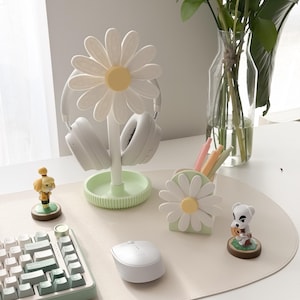 Pu&ograve; includere: Un supporto per cuffie a forma di margherita, un portapenne e delle statuette sono esposti su una scrivania. Il supporto per cuffie &egrave; bianco e verde, con cuffie bianche. Sono visibili anche un mouse e una tastiera bianchi.