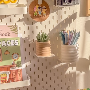 May include: A white pegboard with various items, including a small camera, a corkboard with cartoon figures, a faux succulent, and a pen holder filled with pens. A book titled "Spaces" is visible. The pegboard has multiple holes.