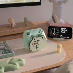 Op de afbeelding: Een lichtgroene standaard voor een gamecontroller met een cartoonkikker die een bloem vasthoudt. De standaard staat op een bureau naast een smartphone met klok en weer, een klein geel figuurtje, een toetsenbord en een bladervormige polssteun.