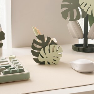 May include: A desk setup featuring a monstera leaf-shaped pen holder in shades of green, holding pens. A matching headset stand and keyboard are also visible. The setup is on a light beige desk mat, with a white mouse.