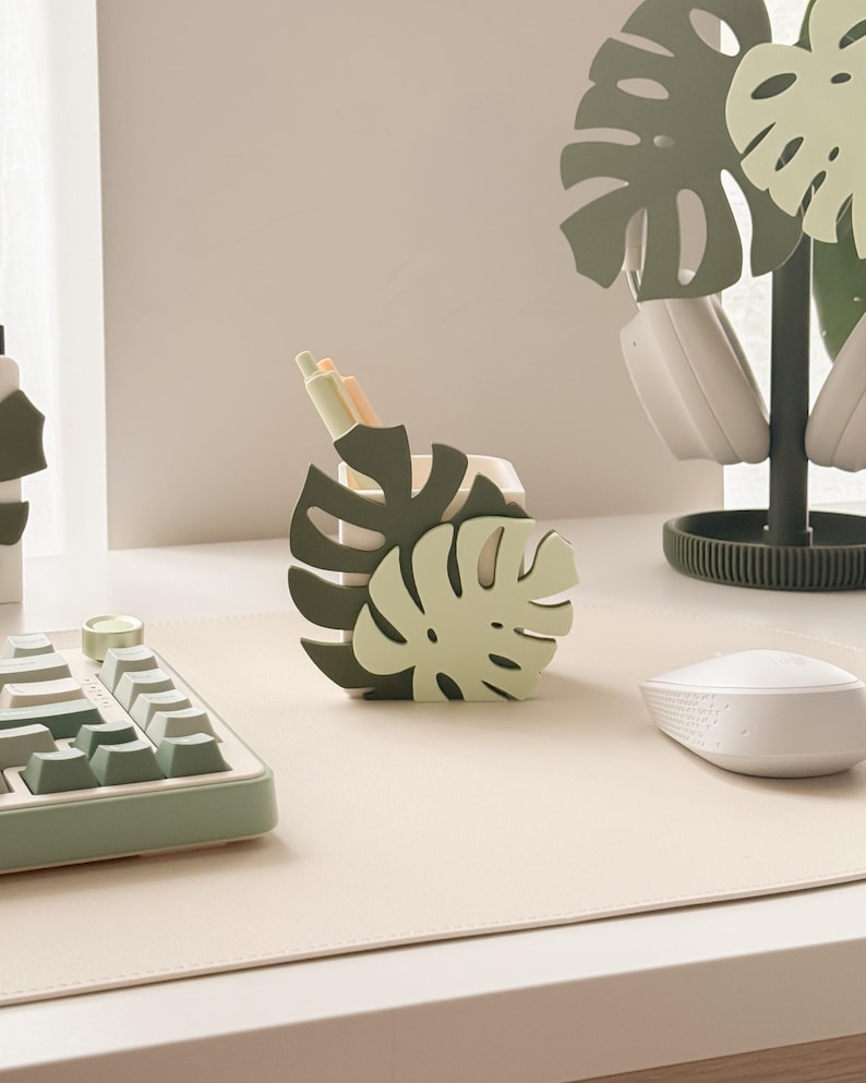 May include: A desk setup featuring a monstera leaf-shaped pen holder in shades of green, holding pens. A matching headset stand and keyboard are also visible. The setup is on a light beige desk mat, with a white mouse.