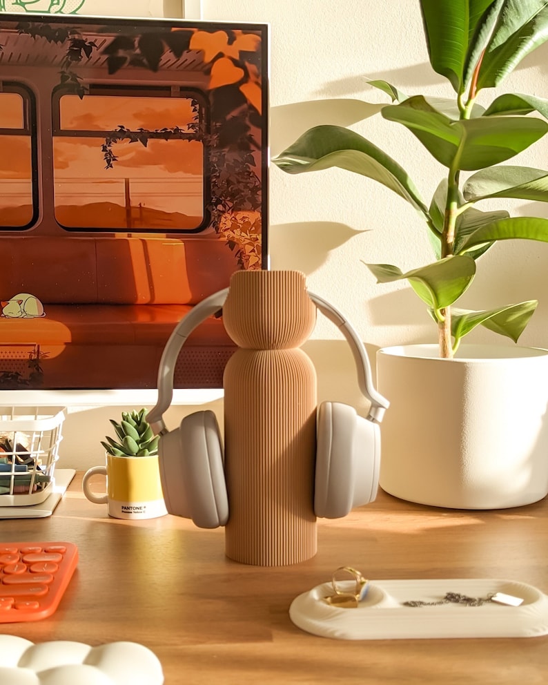 May include: A light brown, ribbed ceramic vase with a pair of grey over-ear headphones resting on it. The headphones are on a wooden desk with a small plant in a yellow mug, a keyboard, and a white tray with a gold ring and a silver chain.