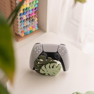 Op de afbeelding: Een grijze en zwarte gamecontroller ligt op een wit oppervlak, met een groene blad-vormige decoratie ervoor. Een verzameling kleurrijke stiften is zichtbaar op de achtergrond, gerangschikt in een bruine organizer. De scène wordt verlicht door natuurlijk licht.