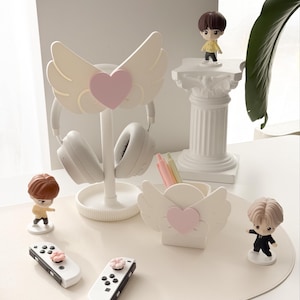 May include: A collection of white desk accessories with a whimsical angel wing and heart design. Includes a headphone stand, pen holder, and two game controllers. Small figurines are also present, along with a pair of white headphones.