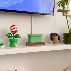 May include: A green and brown video game console stand shaped like a pipe from the Super Mario Bros. video game. The stand is on a white shelf with a green plant, a black video game controller, and a red and green plant toy.