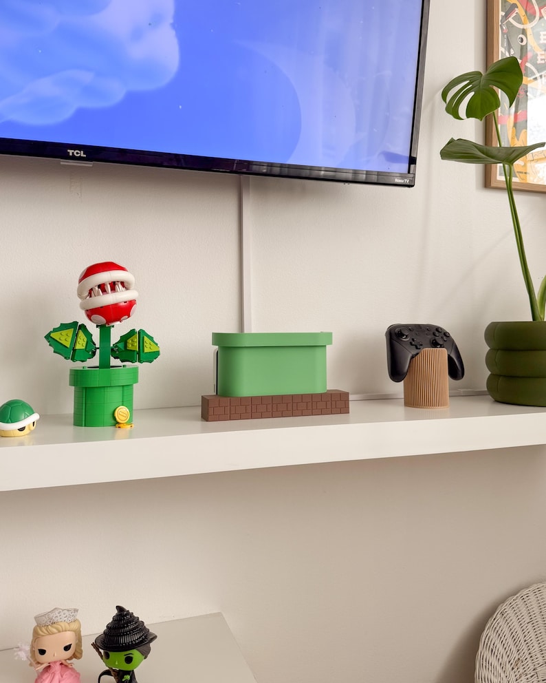 May include: A green and brown video game console stand shaped like a pipe from the Super Mario Bros. video game. The stand is on a white shelf with a green plant, a black video game controller, and a red and green plant toy.