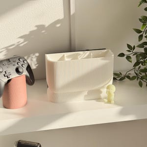 May include: A white, ribbed desk organizer with multiple compartments. A black and gray game controller rests on a pink stand to the left. A small, yellow figurine sits to the right, with a green plant in the background.