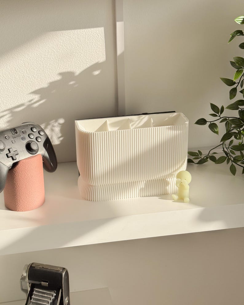 May include: A white, ribbed desk organizer with multiple compartments. A black and gray game controller rests on a pink stand to the left. A small, yellow figurine sits to the right, with a green plant in the background.