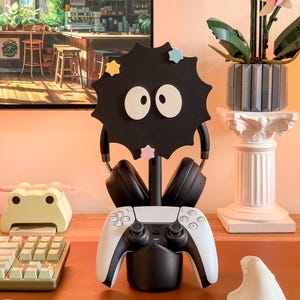 May include: A black headphone stand in the shape of a soot sprite, with white eyes and star accents, holding black headphones and a white and black game controller. A keyboard and a frog-shaped desk tidy are also visible.