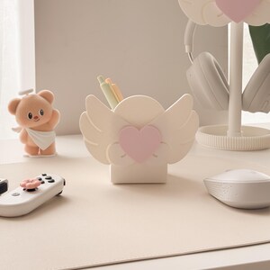 May include: A desk setup featuring a white angel wing-shaped pen holder with a pink heart, a matching headphone stand, and a teddy bear figurine. The desk also has a white mouse, a game controller, and a beige desk mat.