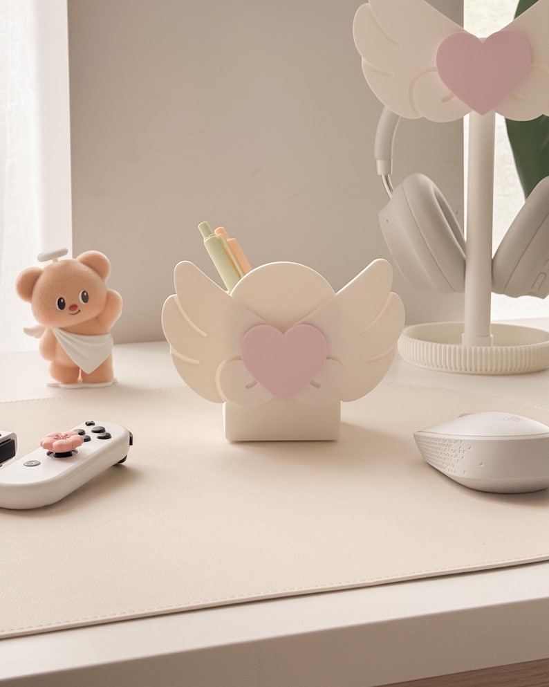 May include: A desk setup featuring a white angel wing-shaped pen holder with a pink heart, a matching headphone stand, and a teddy bear figurine. The desk also has a white mouse, a game controller, and a beige desk mat.