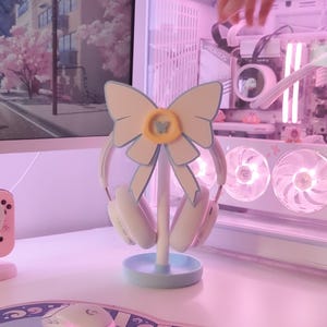 May include: A light blue and white headset stand with a bow design. The stand has a butterfly charm and is holding a pair of white headphones.