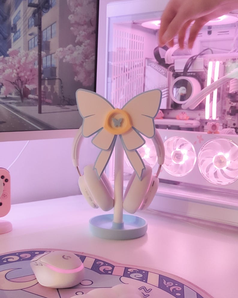 May include: A light blue and white headset stand with a bow design. The stand has a butterfly charm and is holding a pair of white headphones.