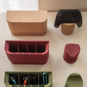 May include: A collection of modern, minimalist gaming accessories in various colors. Includes a black game controller, storage boxes, and stands in shades of beige, red, and green. The accessories are designed to organize and display gaming equipment.