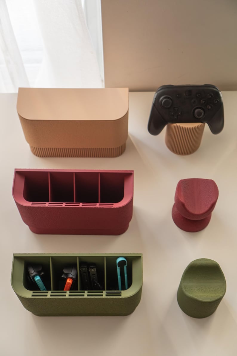 May include: A collection of modern, minimalist gaming accessories in various colors. Includes a black game controller, storage boxes, and stands in shades of beige, red, and green. The accessories are designed to organize and display gaming equipment.