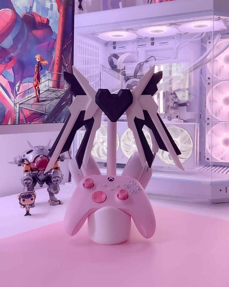 May include: A pink and white gaming controller stand with a black heart design. The stand has two wings that extend out from the sides. The controller is a pink Xbox controller.