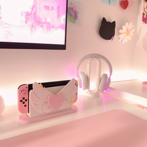 May include: A pink and white Nintendo Switch console stand with angel wings and a heart. The stand is on a pink desk with a white headset on a stand.