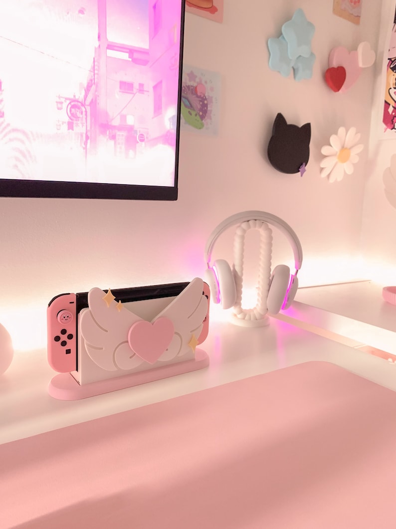 May include: A pink and white Nintendo Switch console stand with angel wings and a heart. The stand is on a pink desk with a white headset on a stand.