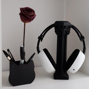May include: A black cat-shaped pen holder with pens and markers, a dried burgundy rose, and a black headphone stand holding white and black headphones. The items are on a white shelf.