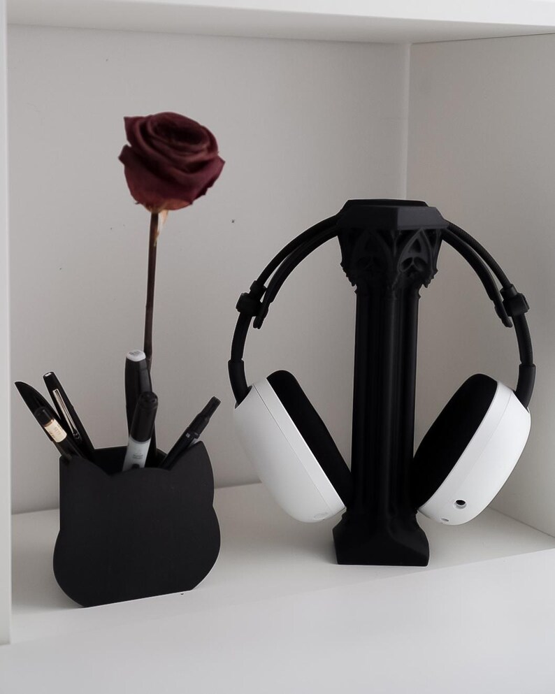 May include: A black cat-shaped pen holder with pens and markers, a dried burgundy rose, and a black headphone stand holding white and black headphones. The items are on a white shelf.