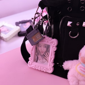 May include: Black shoulder bag with silver hardware, a pink framed photo charm, and a gray game cartridge charm. The pink framed photo charm has a picture of a person. A small pink plush toy is also visible.