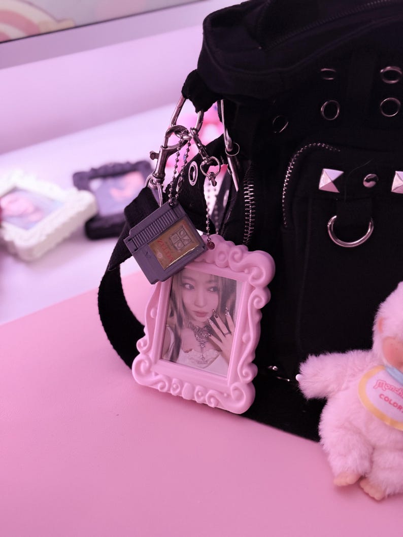 May include: Black shoulder bag with silver hardware, a pink framed photo charm, and a gray game cartridge charm. The pink framed photo charm has a picture of a person. A small pink plush toy is also visible.