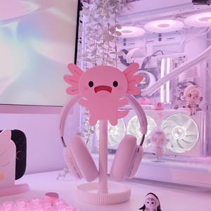 May include: A pink axolotl-shaped headphone stand holding white headphones. The stand is on a white desk with a small penguin figurine and other decorative items. The background includes a computer and hanging plants.