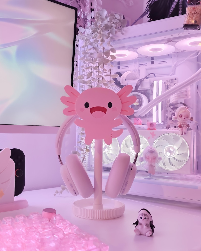 May include: A pink axolotl-shaped headphone stand holding white headphones. The stand is on a white desk with a small penguin figurine and other decorative items. The background includes a computer and hanging plants.