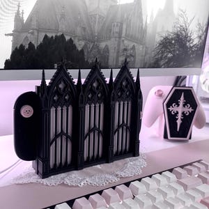 Può includere: Una base di ricarica nera per console di gioco con un design a tre pannelli ispirato alle finestre delle cattedrali gotiche, caratterizzato da archi a sesto acuto e trafori complessi. Poggia su un centrino bianco accanto a un controller rosa.