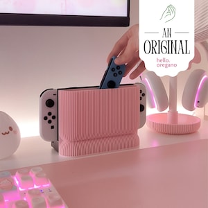 Op de afbeelding: Een roze geribbelde consolehouder met een blauwe controller die wordt geplaatst. De houder is ontworpen om een gameconsole en controllers te bevatten. De afbeelding toont ook een witte en roze gaming-opstelling met een toetsenbord en een koptelefoon.