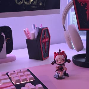 May include: A black coffin-shaped pen holder with red trim and a cross design, filled with pens and pencils. A figurine of a doll with red hair and a keyboard are on a pink desk. A headset and a decorative coffin are also visible.