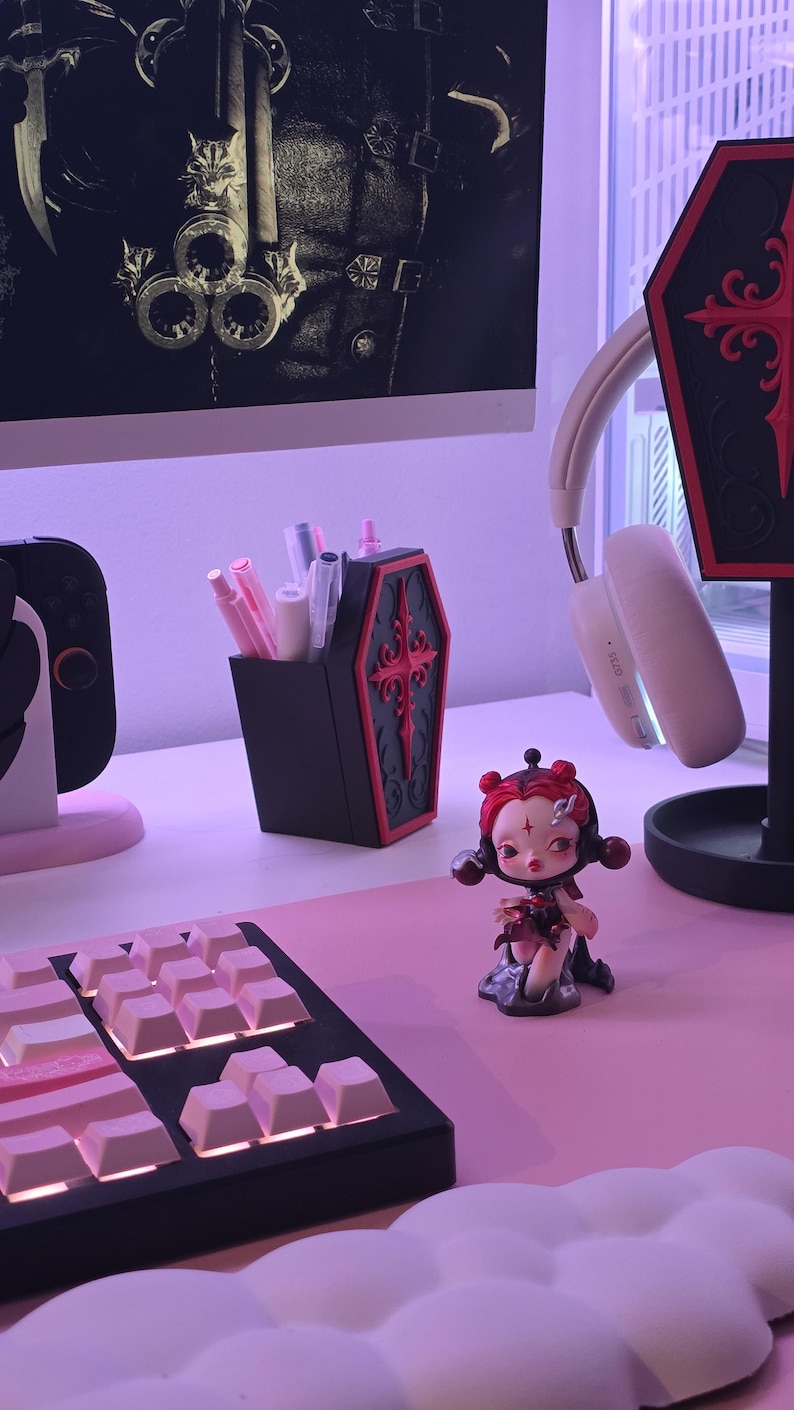 May include: A black coffin-shaped pen holder with red trim and a cross design, filled with pens and pencils. A figurine of a doll with red hair and a keyboard are on a pink desk. A headset and a decorative coffin are also visible.