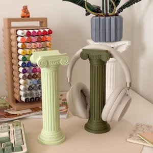 Puede incluir: Dos columnas decorativas, una verde claro y otra verde oliva. Un par de auriculares blancos descansa sobre la columna verde oliva. Tambi&eacute;n se ven un portal&aacute;pices marr&oacute;n y un teclado.