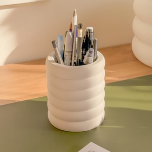 May include: A cream-colored, cylindrical pen holder with a ridged design, filled with various pens, pencils, and markers. The holder sits on a green surface, suggesting a desk or workspace. The pens are of different colors and sizes.