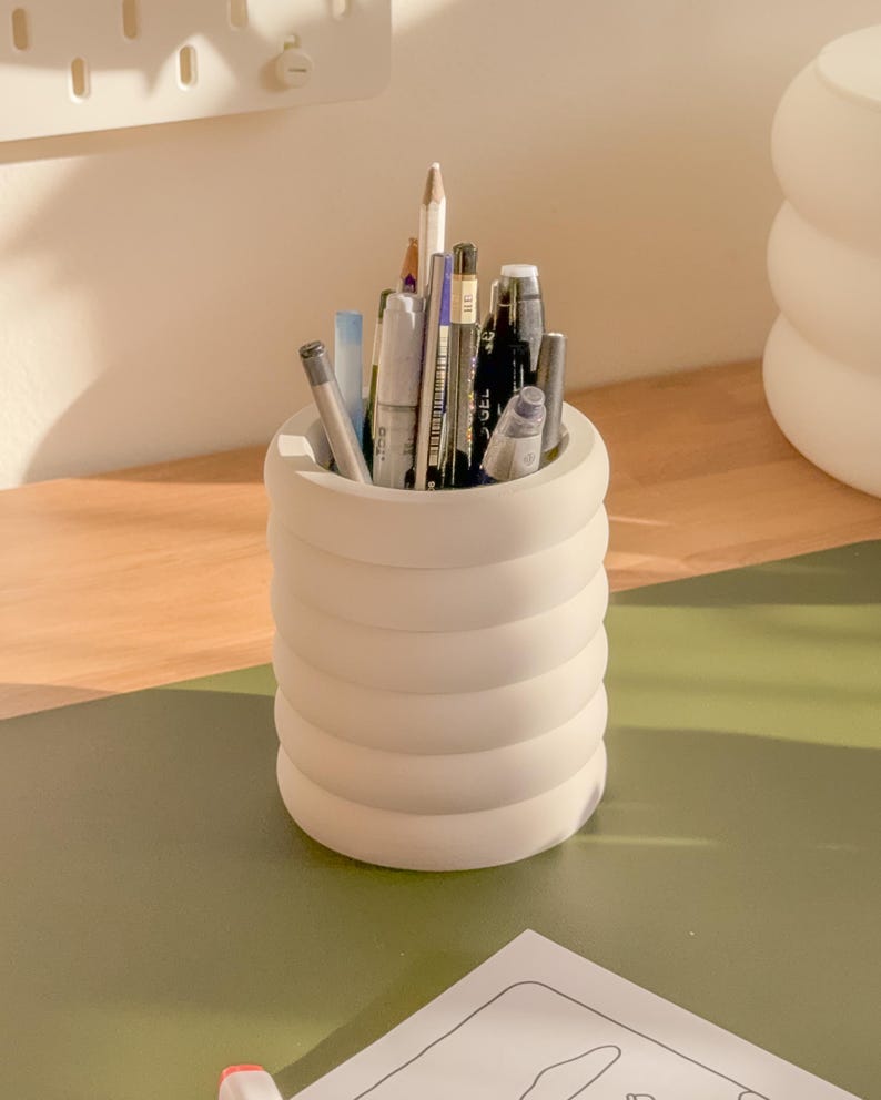 May include: A cream-colored, cylindrical pen holder with a ridged design, filled with various pens, pencils, and markers. The holder sits on a green surface, suggesting a desk or workspace. The pens are of different colors and sizes.