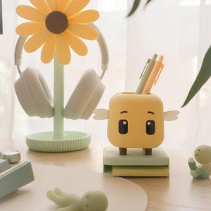 Puede incluir: Una composición de escritorio con un portalápices amarillo con forma de personaje de dibujos animados, bolígrafos, un soporte para auriculares con forma de margarita con auriculares blancos y pequeñas figuras. La paleta de colores incluye amarillo, verde y blanco.