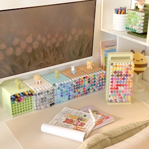 May include: A collection of desk organizers holding numerous colored markers, featuring a vertical stand and several horizontal grid-style containers topped with small figurines. A coloring book with a partially colored illustration sits on the desk.
