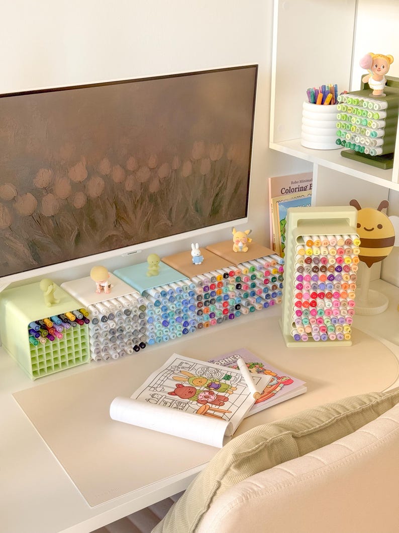 May include: A collection of desk organizers holding numerous colored markers, featuring a vertical stand and several horizontal grid-style containers topped with small figurines. A coloring book with a partially colored illustration sits on the desk.