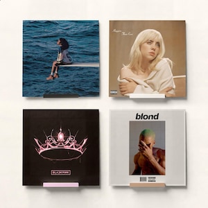 May include: Four square album art prints displayed on a white wall. The prints feature album covers, including one with a person sitting on a diving board over water, another with a blonde woman, a black cover with a pink crown, and a cover with the word "blond".