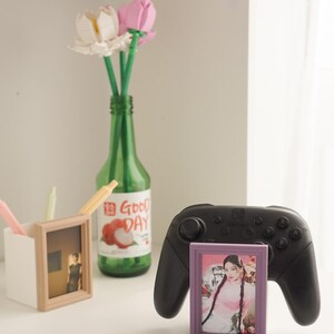 May include: A still life arrangement featuring a green bottle with "Good Day" text, holding artificial flowers. A black game controller and two small picture frames are also present, one with a photo of a person.