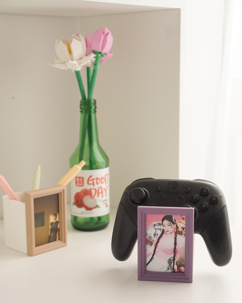 May include: A still life arrangement featuring a green bottle with "Good Day" text, holding artificial flowers. A black game controller and two small picture frames are also present, one with a photo of a person.