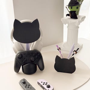 May include: A black cat-shaped desk organizer holds a black game controller and a pair of headphones. A matching pen holder with pens and a witch figurine are also on the desk. Two game controllers are in the foreground.