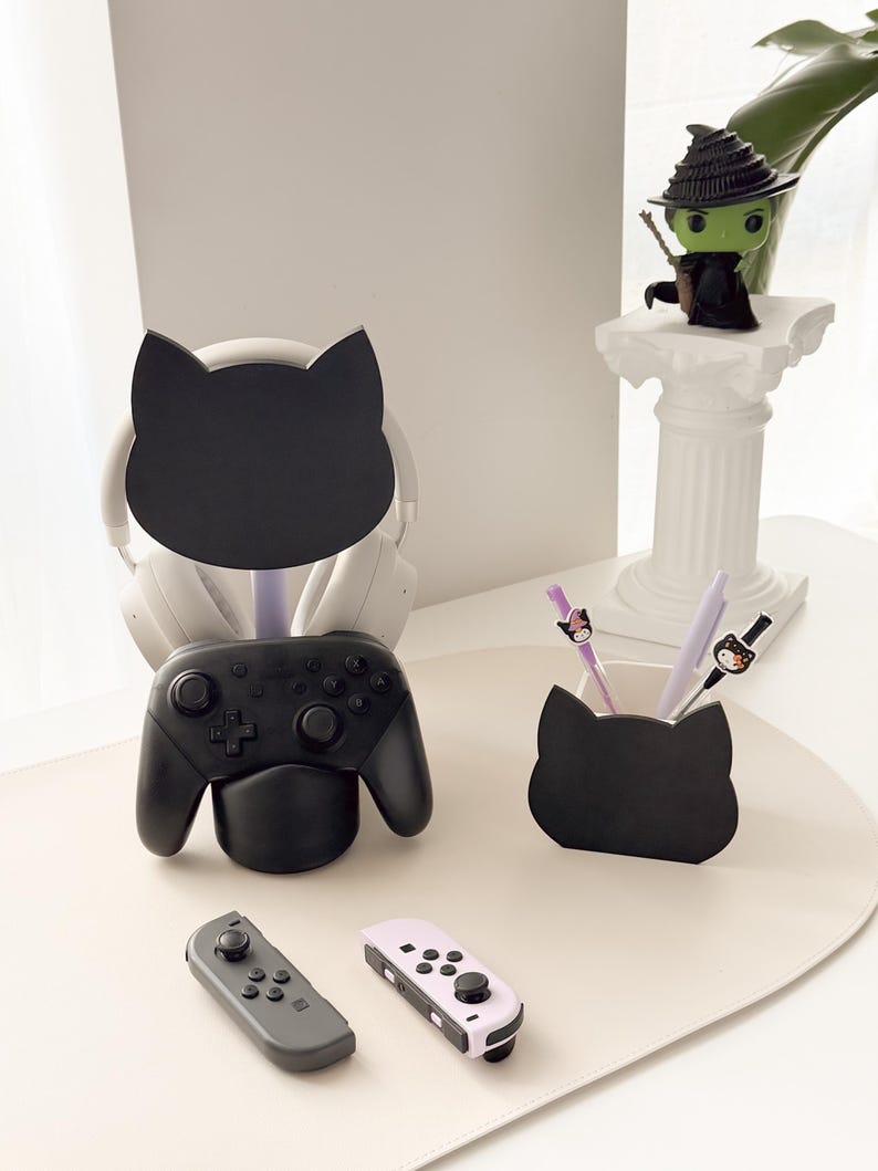 May include: A black cat-shaped desk organizer holds a black game controller and a pair of headphones. A matching pen holder with pens and a witch figurine are also on the desk. Two game controllers are in the foreground.
