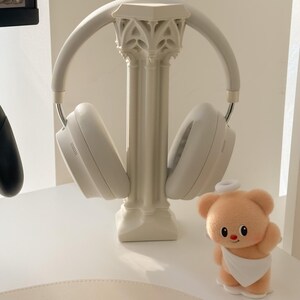 May include: White wireless headphones resting on a decorative column-shaped stand. A small, plush teddy bear with a halo and a white cloth is positioned nearby. The scene is set on a white surface.