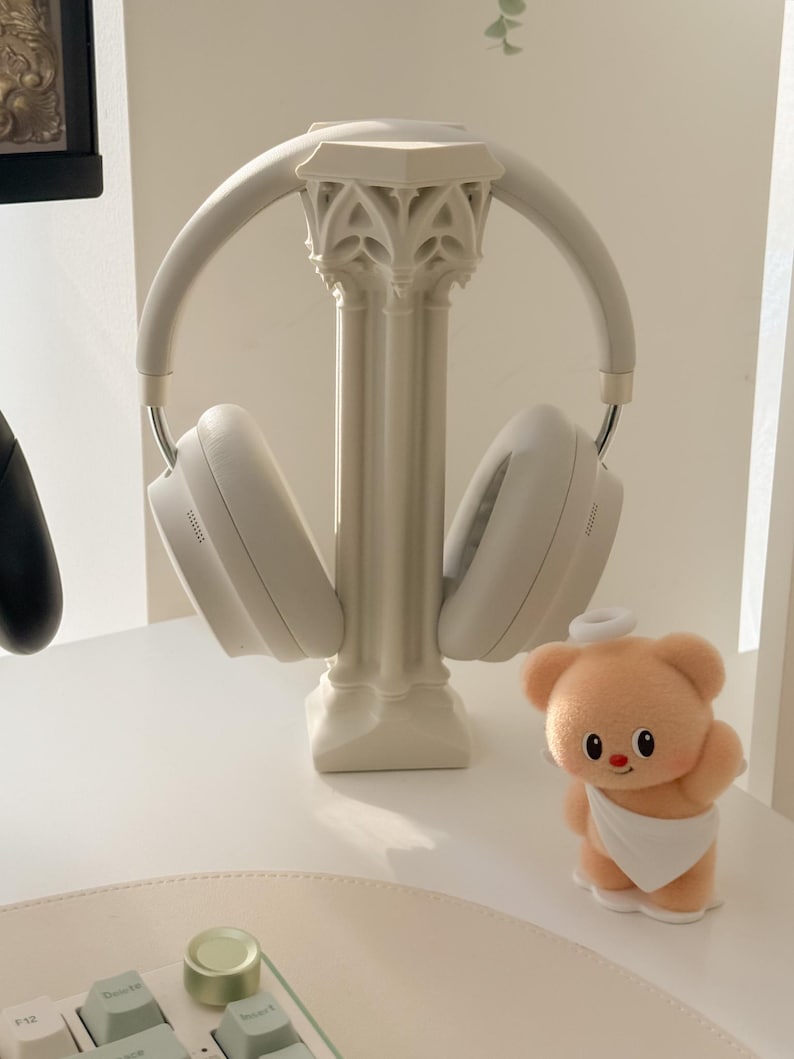 May include: White wireless headphones resting on a decorative column-shaped stand. A small, plush teddy bear with a halo and a white cloth is positioned nearby. The scene is set on a white surface.