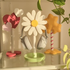 May include: A white daisy-shaped headphone stand with a pair of gray headphones hanging from it. The stand is on a white surface with a pink heart-shaped stand and a yellow and orange tree-shaped stand.