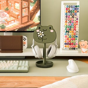 May include: A green frog-shaped headphone stand with a daisy design, holding white headphones. The stand is on a desk with a keyboard, mouse, and a marker holder. A computer monitor displays a cozy cabin scene.